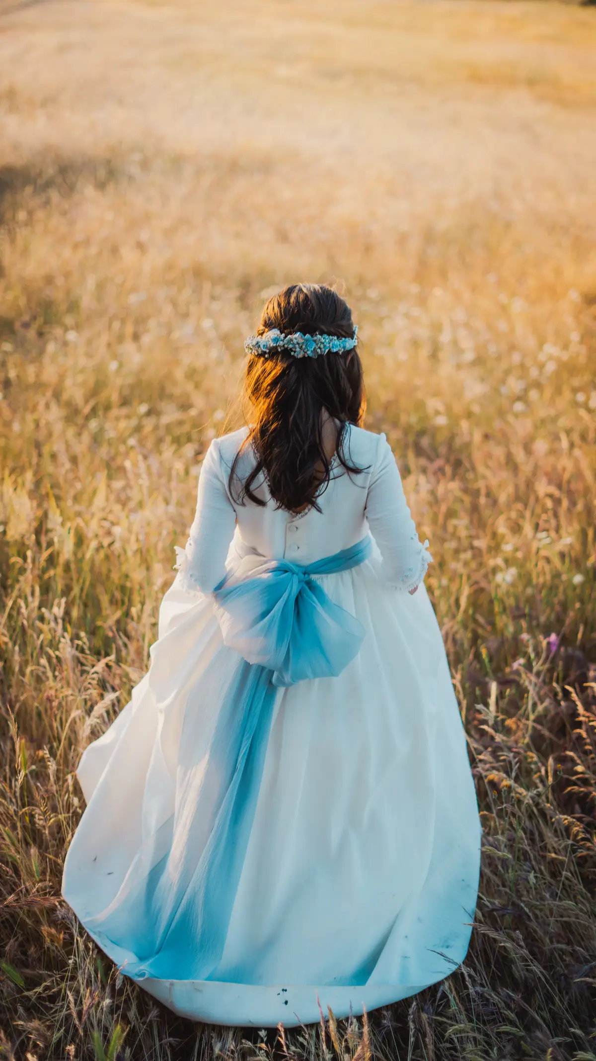 Niña de comunión caminando de espaldas entre hierbas iluminadas por el atardecer