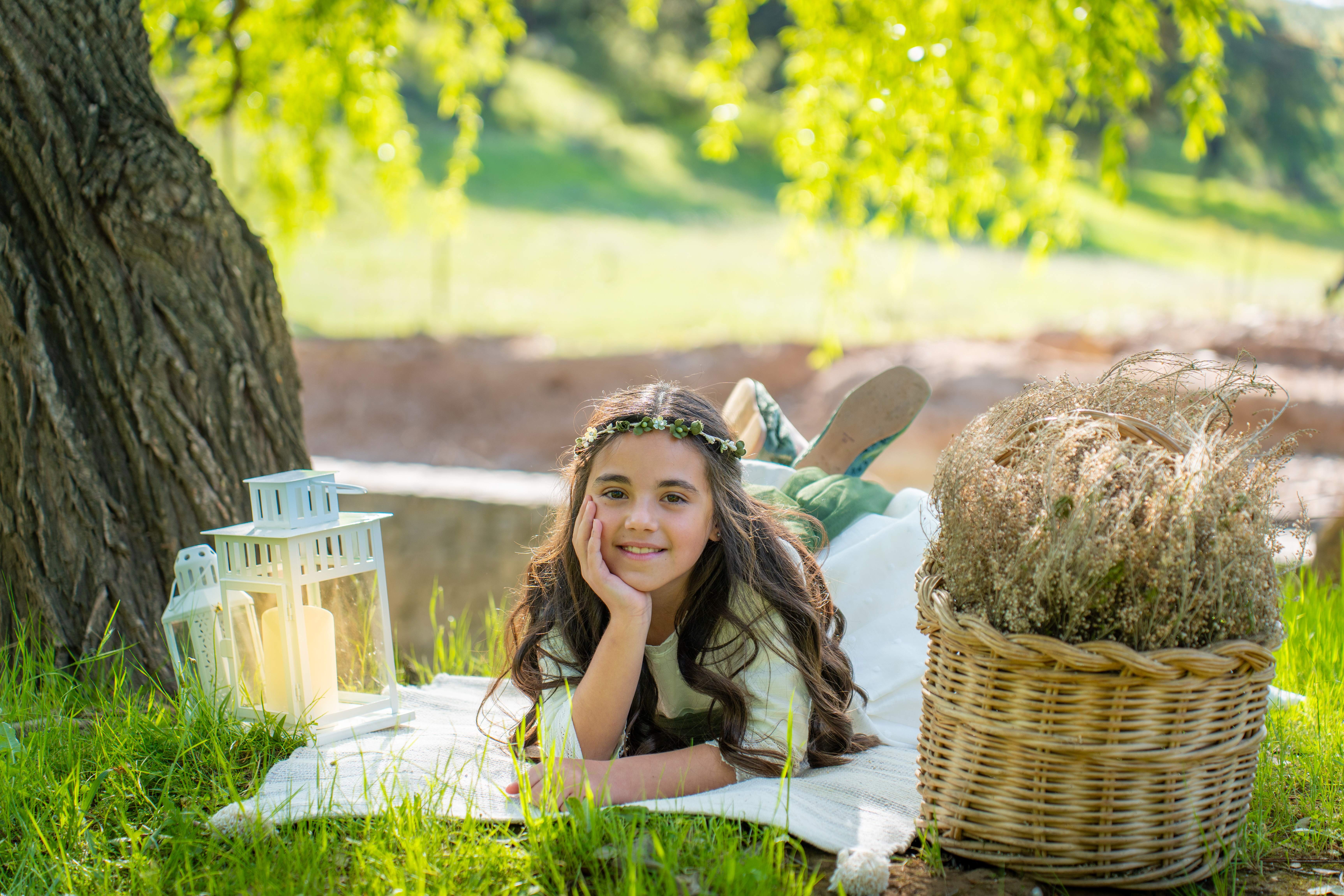 Niña de comunión tumbada sobre una manta junto a un árbol y una cesta de flores