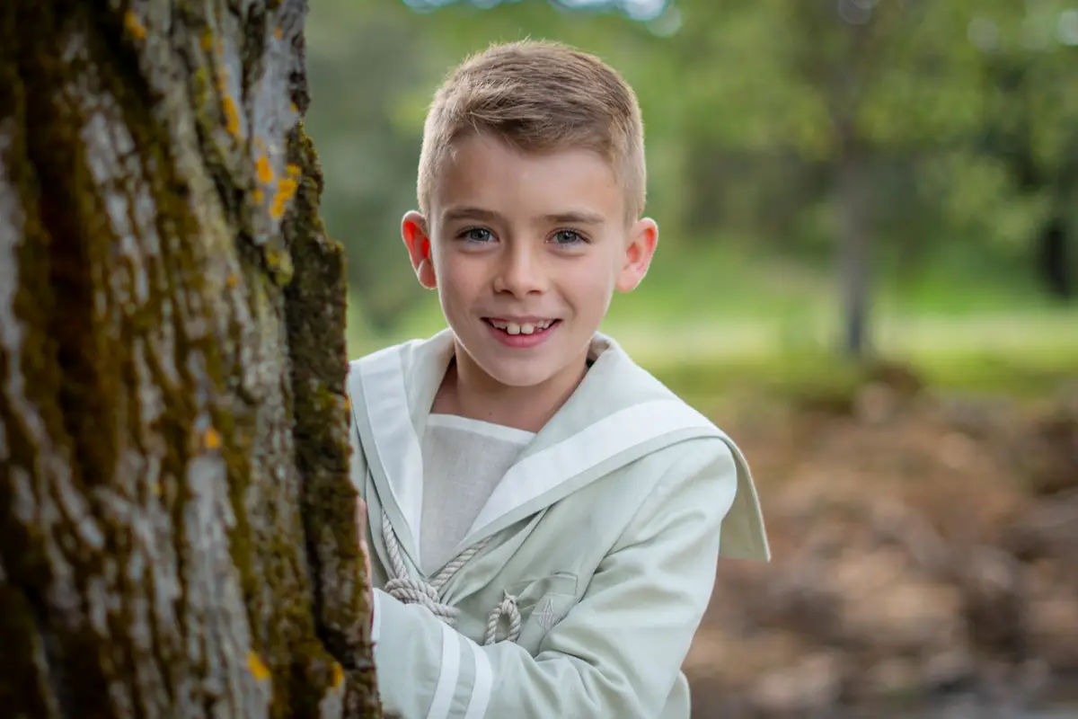 Niño vestido de primera comunión sonriendo junto al tronco de un árbol