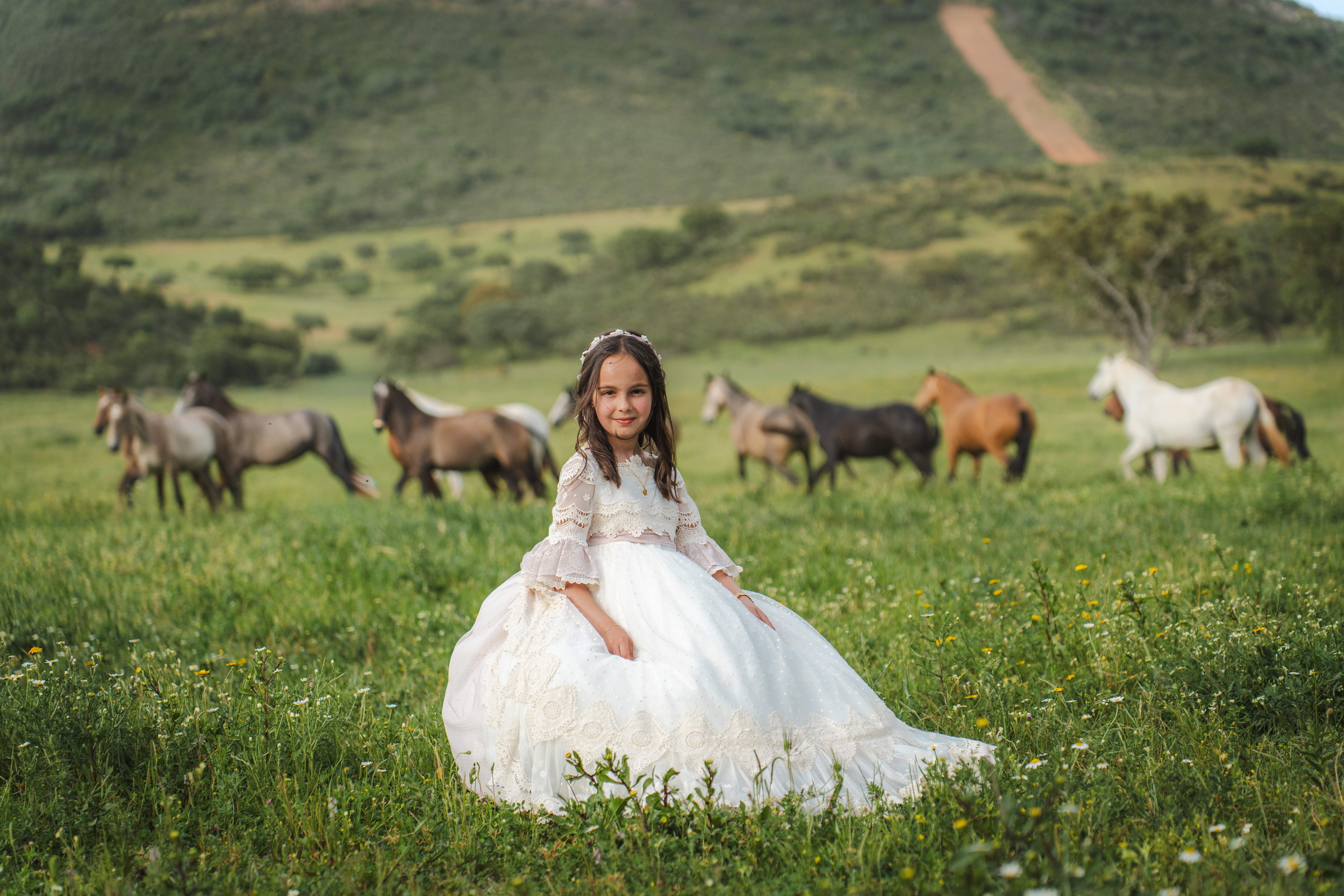 Niña con vestido de comunión sentada en un prado con caballos al fondo, en Fuenlabrada de los Montes (Badajoz)